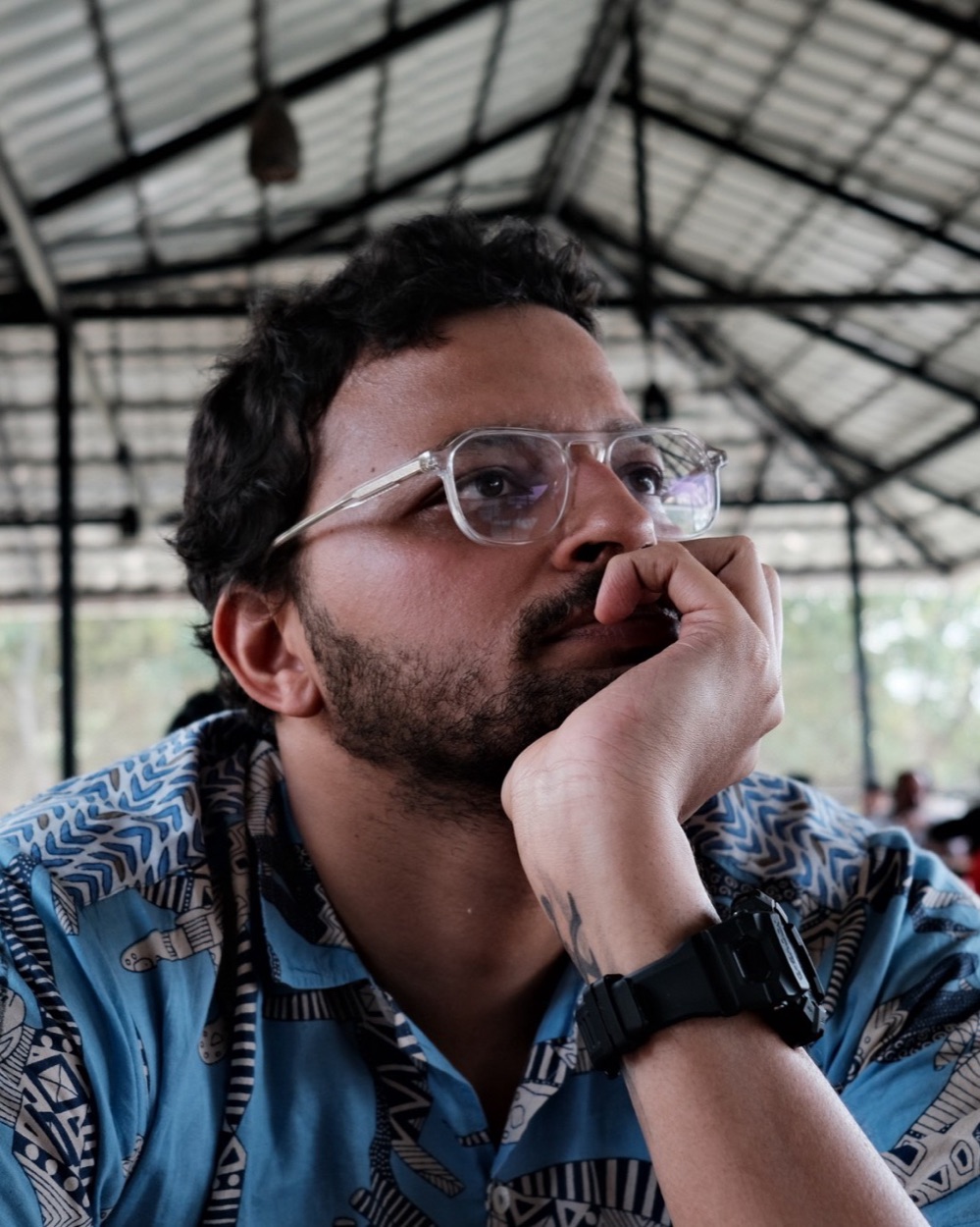 Portrait of Aakash Karkare, chin resting on his hand, wearing clear-framed glasses and a blue printed shirt
