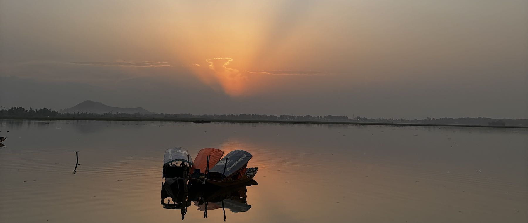 Sunset over Dal Lake: rays fan out from behind clouds above distant hills, and a cluster of covered shikaras sits on the still water with its reflection