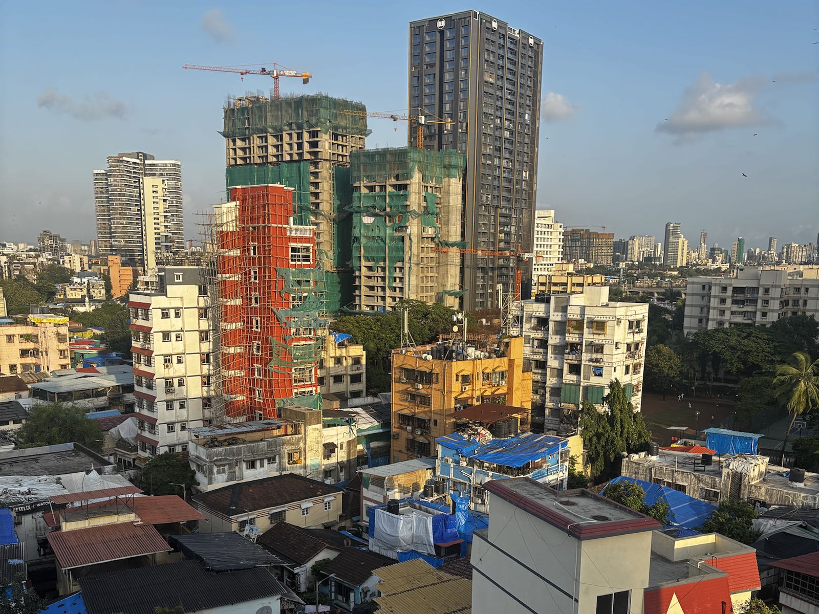 View over Mumbai rooftops: towers wrapped in green and orange scaffolding rise behind old tiled roofs and blue tarpaulins, with cranes against an evening sky