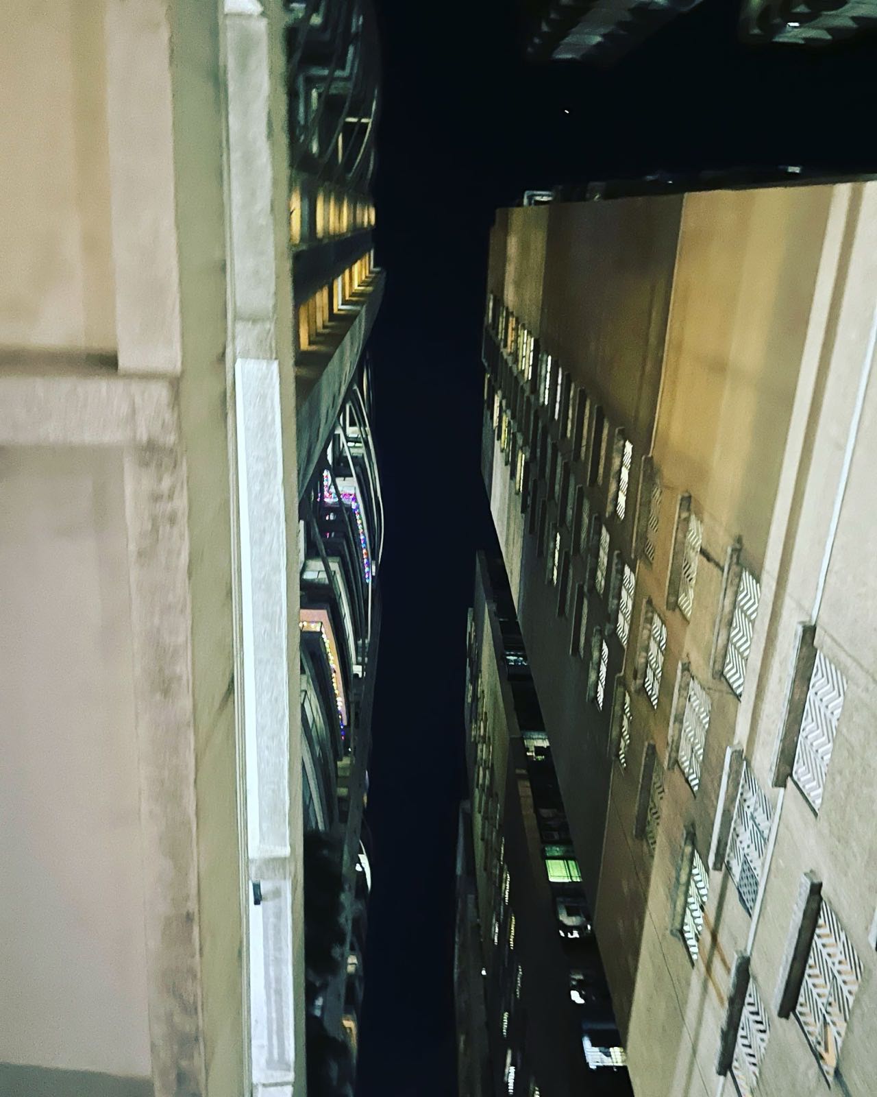 Looking straight up at night between tower blocks, their lit windows and grilles closing in on a narrow strip of dark sky