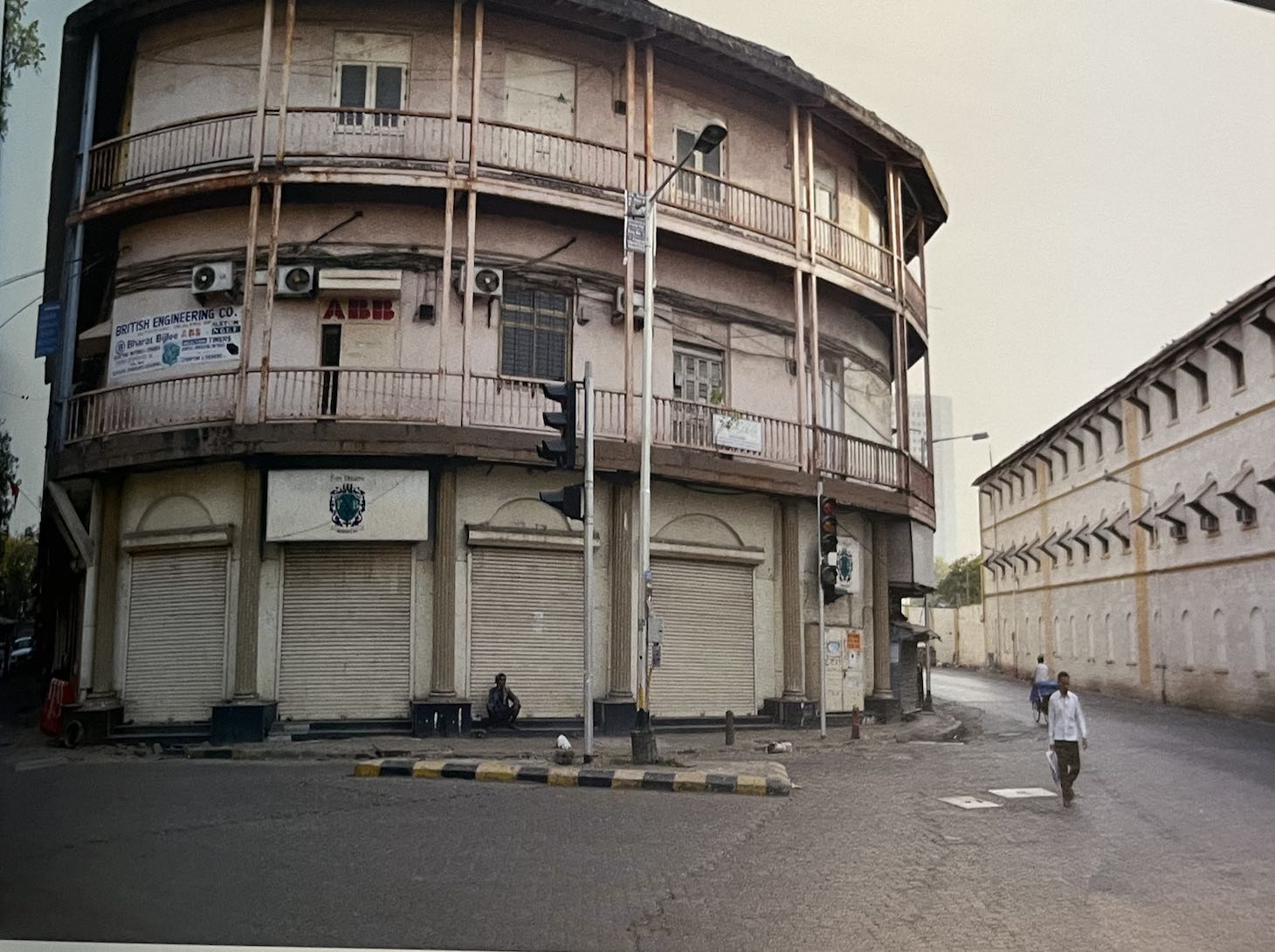 A curved three-storey corner building in Fort, Mumbai, with wraparound balconies above shuttered shops, a man walking along the empty street beside it